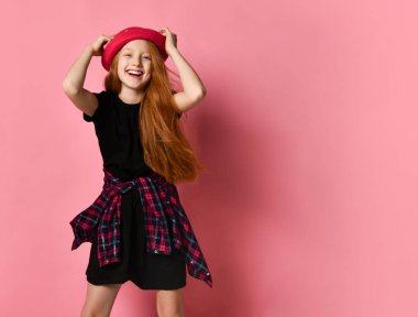 Siyah elbiseli, belinde kareli gömlek olan genç bir çocuk. Gülümsüyor, kırmızı şapkasına dokunuyor, pembe arka planda poz veriyor. Kapat.