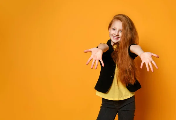 Ingefära tonåring i svart jacka och byxor, gul t-shirt. Hon ler, sträcker sig efter dig, poserar på orange bakgrund. Närbild — Stockfoto