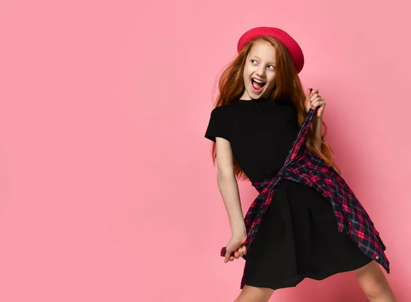 Niño adolescente con vestido negro y sombrero rojo. Ella aprieta la camisa a cuadros en su cintura mientras posa sobre el fondo rosa. De cerca. —  Fotos de Stock