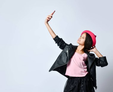 Siyah deri ceketli, etekli ve pembe şapkalı genç. Akıllı telefon kullanarak selfie çekiyor, beyaza izole edilmiş poz veriyor. Kapat.