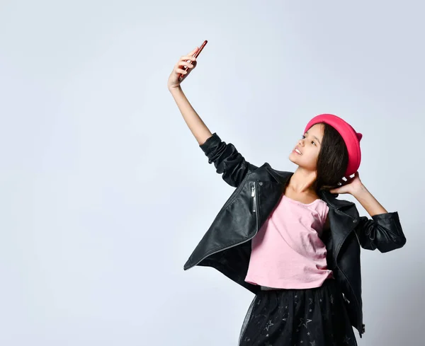 Enfant adolescent en veste en cuir noir, jupe et chapeau rose. Elle prend selfie à l'aide d'un smartphone, posant isolée sur blanc. Gros plan — Photo