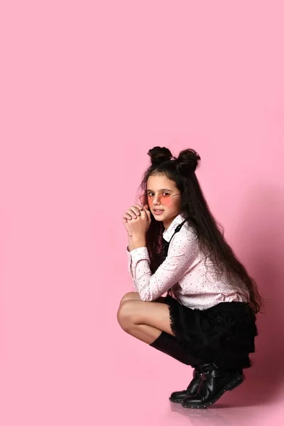 Niño adolescente con el pelo largo, en gafas de sol, blusa blanca, falda negra y botas. Ella posando en cuclillas sobre fondo rosa. De cerca. — Foto de Stock