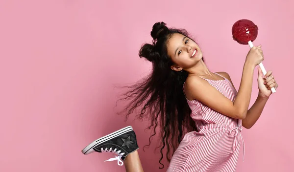 Tiener kind met chique kapsel, in gestreepte jurk. Ze heeft een grote rode lolly, poserend op een roze achtergrond. Sluiten. — Stockfoto