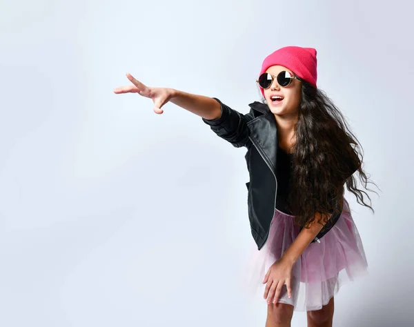 Adolescente en chaqueta de cuero negro, falda, sombrero rosa y gafas de sol. Ella sonrió mientras posaba de pie aislada sobre blanco. De cerca. — Foto de Stock