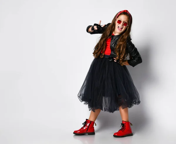 Joven chica positiva más tamaño en ropa de estilo rock brillante, botas rojas y gafas de pie y mostrando signo de paz con los dedos — Foto de Stock