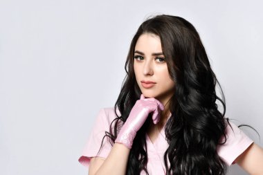 Attractive woman in pink uniform and disposable gloves posing on white. She is thinking of something. Medical staff, beautician, tattooist, hairdresser.