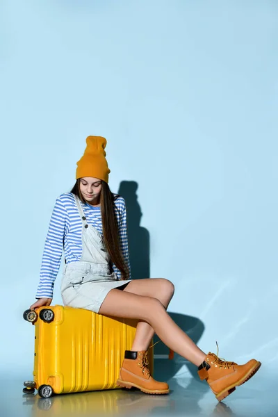 Teenager in jeans overall skirt, striped sweatshirt, boots and hat. She sitting on yellow suitcase, blue background. Full length — 스톡 사진