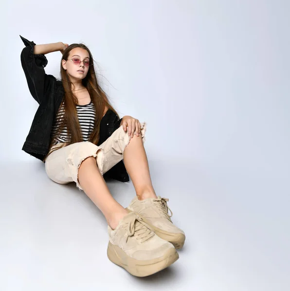 Tiener dame met zonnebril, gestreept t-shirt, zwarte denim jas, broek en sneakers. Ze zit op de vloer geïsoleerd op wit. Sluiten. — Stockfoto