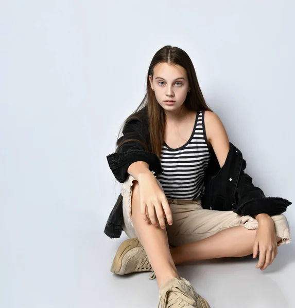 Adolescente en camiseta a rayas, chaqueta vaquera negra, pantalones y zapatillas de deporte. Ella está sentada en el suelo aislada de blanco. De cerca. —  Fotos de Stock