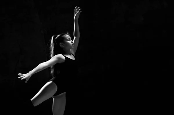 Jovem ginasta menina em corpo esporte preto e uppers especiais de pé e fazendo pose ginástica sobre fundo escuro — Fotografia de Stock