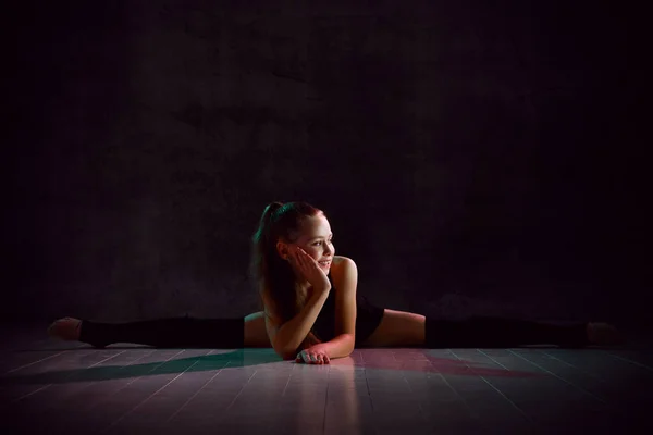 Young smiling girl gymnast in black sport body and uppers sitting in twine on floor and holding pink gymnastic ball in hands — ストック写真