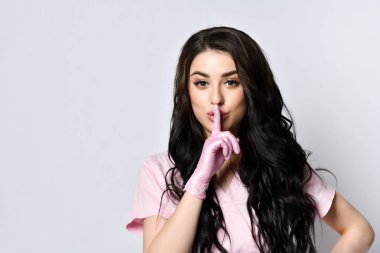 Pembe üniformalı ve tek kullanımlık eldivenli genç bir kadın beyaz üzerine izole edilmiş poz veriyor. Sessizlik işareti gösteriyor. Uzayı kopyala Kapat.