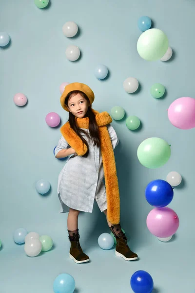 Klein brunette aziatisch meisje in oversized shirt jurk, bruin baret en sjaal, Ze poseren met ballonnen op blauwe achtergrond. — Stockfoto