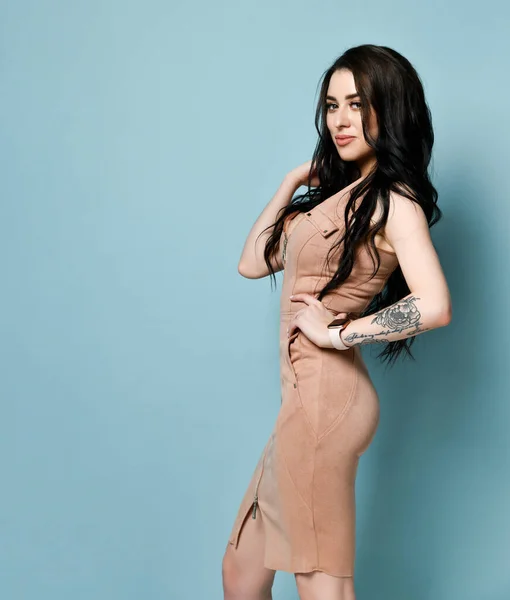 Brunett modell i beige klänning. Det är klocka på hennes tatuerade hand. Stående i sidled mot blå studio bakgrund. Närbild — Stockfoto