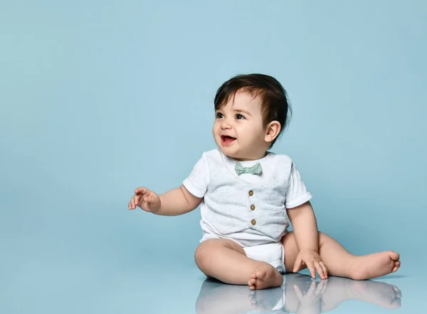 Ett litet barn i vit kroppsdräkt som en väst med fluga, barfota. Han sitter på golvet mot blå bakgrund. Närbild — Stockfoto