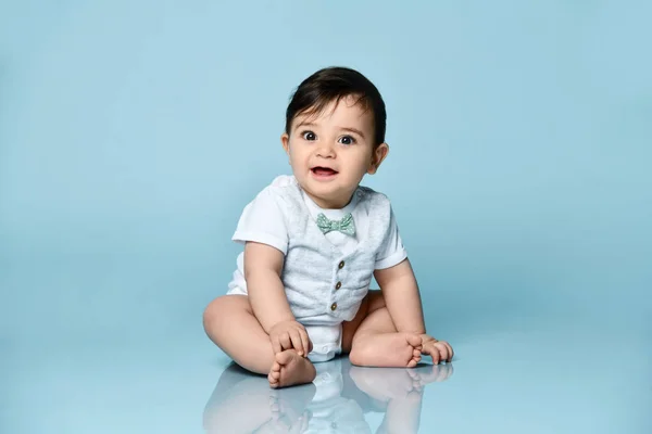 Bébé garçon en body blanc en gilet avec noeud papillon, pieds nus. Il sourit, assis par terre sur fond bleu. Gros plan — Photo