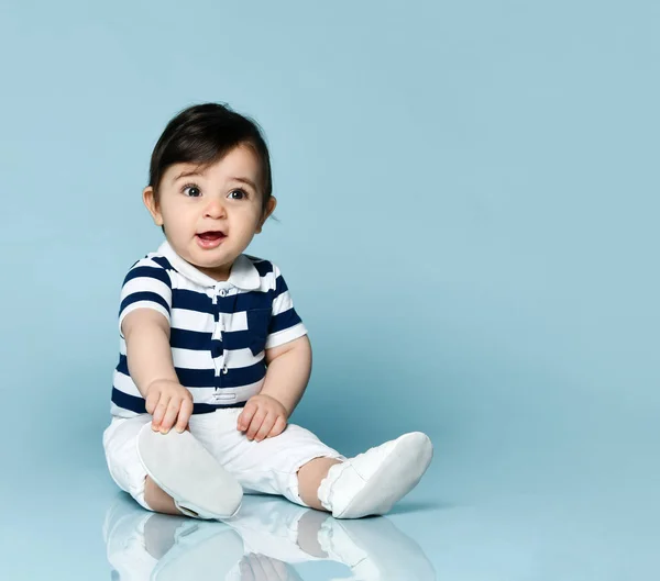 Bébé garçon en t-shirt rayé, pantalon blanc et chaussons. Il sourit, assis sur le sol sur fond bleu studio. Gros plan — Photo