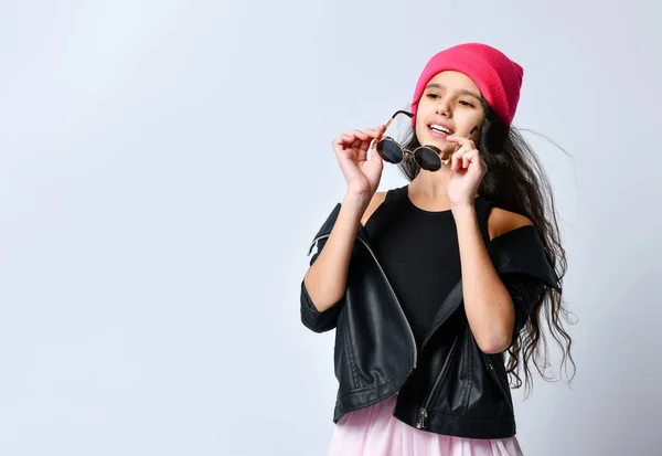 Teenage child in black leather jacket, skirt and pink hat. She is holding her sunglasses, posing isolated on white. Close up — 스톡 사진