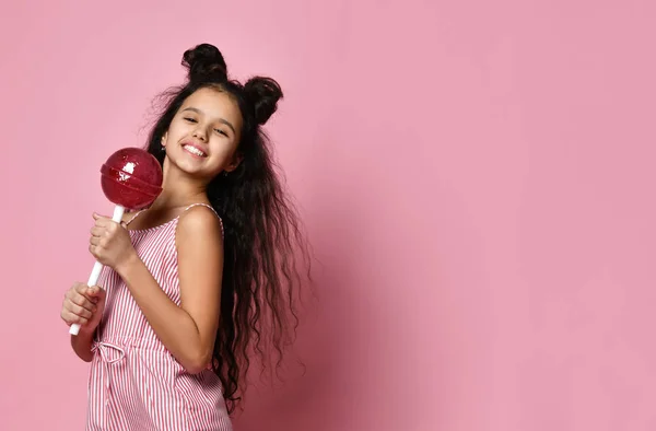 Tiener met mooi kapsel, in gestreepte jurk. Ze houdt een enorme rode lolly vast, poserend op een roze achtergrond. Sluiten. — Stockfoto