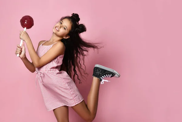 Teenage child with fancy hairstyle, in striped dress. She is holding a big red lollipop, posing on pink background. Close up — 图库照片