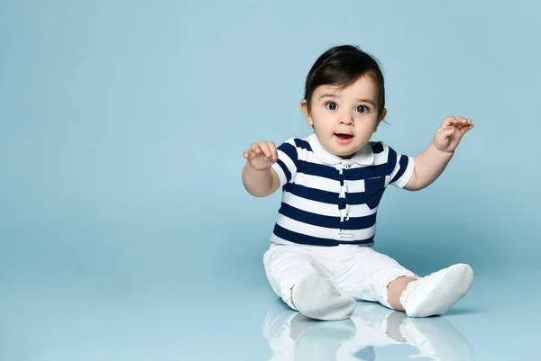 Baby boy in striped t-shirt, white pants and booties. He is smiling, sitting on floor against blue studio background. Close up — Stock Photo, Image