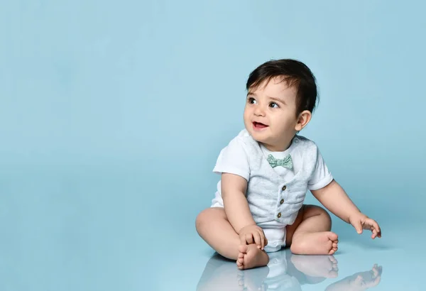 Bébé garçon en body blanc en gilet avec noeud papillon, pieds nus. Il sourit, assis par terre sur fond bleu. Gros plan — Photo