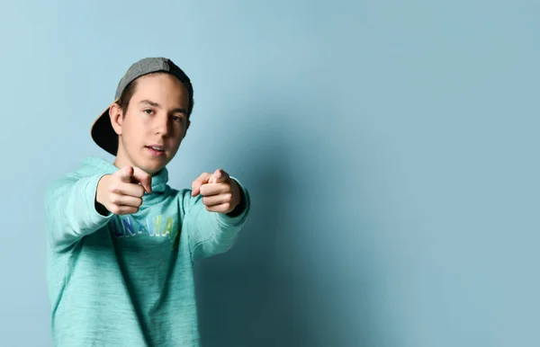 Joven adolescente positivo en sudadera con capucha azul de pie y apuntando a la cámara con los dedos índice sobre fondo de pared azul pastel —  Fotos de Stock