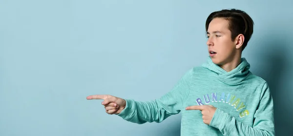 Joven adolescente con capucha azul de pie y señalando a un lado algo con los dedos sobre fondo de pared azul pastel —  Fotos de Stock