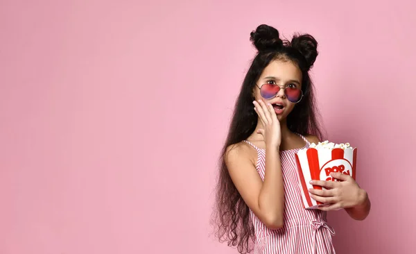 Teenage child in striped dress and sunglasses. She looking wondered, holding pack of popcorn, posing on pink background. Close up — 图库照片