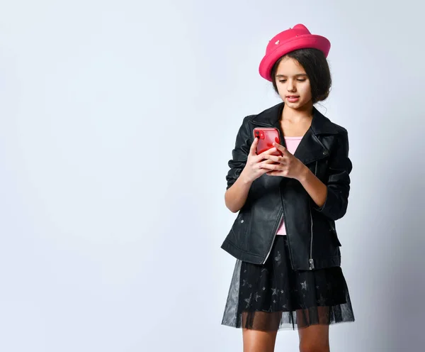 Jeune fille en cuir noir veste, jupe, t-shirt et chapeau rose. Elle utilise un smartphone, posant isolée sur du blanc. Gros plan — Photo