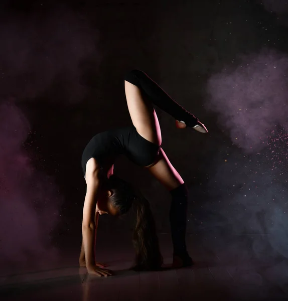 Młoda gimnastyczka w czarnym sporcie ciała i specjalne obuwie stojące w pozycji mostu i utrzymanie równowagi na ciemnym tle — Zdjęcie stockowe
