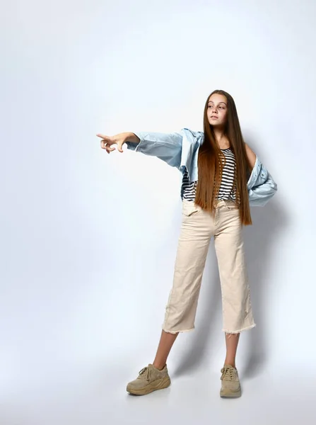 Tienervrouw in gestreept t-shirt, sneakers, spijkerjasje en broek. Richt op iets, poseren geïsoleerd op wit. Totale lengte — Stockfoto