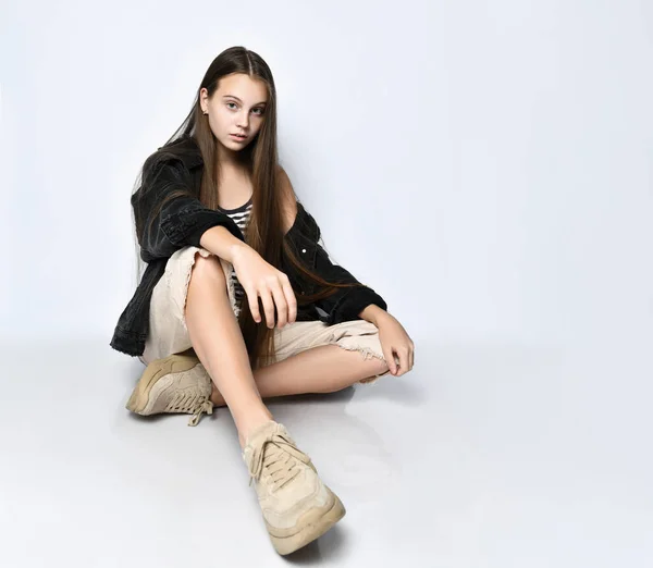 ストライプTシャツ、ブラックデニムジャケット、パンツ、スニーカーのティーンエイジャー。彼女は白い床の上に座っている。閉じろ! — ストック写真