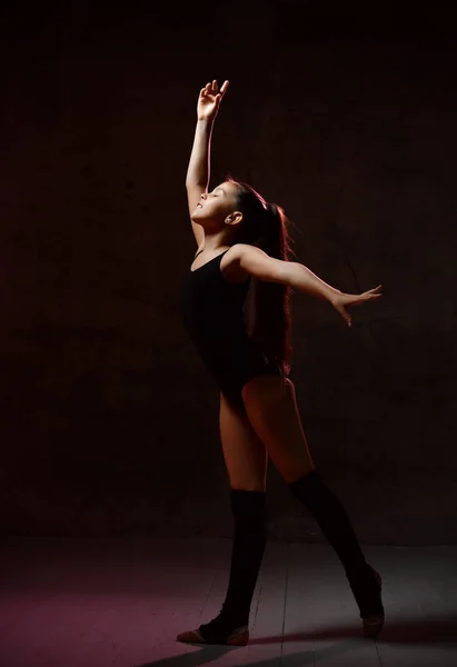 Ung flicka gymnast i svart sport kropp och speciella överdelar stående och göra gymnastiska poser över mörk bakgrund — Stockfoto