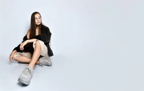 Adolescente em camiseta listrada, casaco de ganga preta, calças e tênis. Ela está sentada no chão isolada em branco. Fechar — Fotografia de Stock