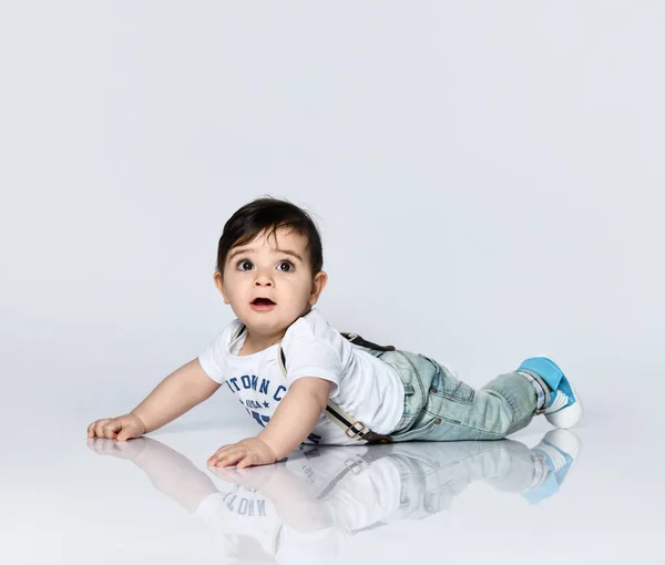 碑文付きのTシャツの小さな幼児、サスペンダー付きのジーンズ。彼は白い背景に隔離された床に横たわっている。閉じろ! — ストック写真
