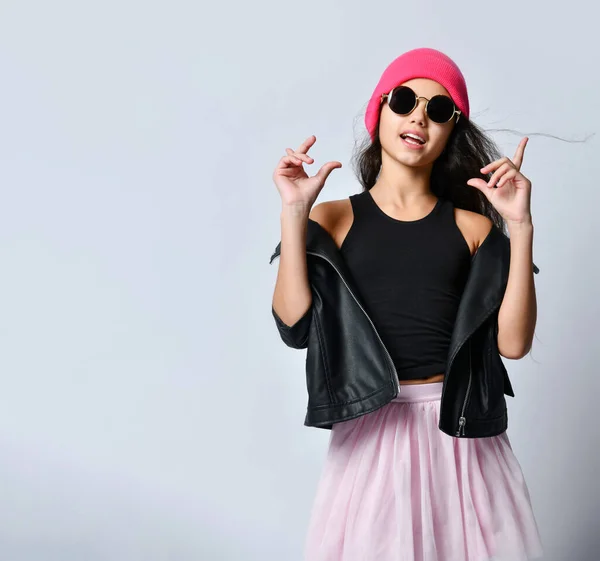 Teenage femelle en cuir noir veste chapeau rose et lunettes de soleil. Elle gesticule en posant isolée sur du blanc. Gros plan — Photo