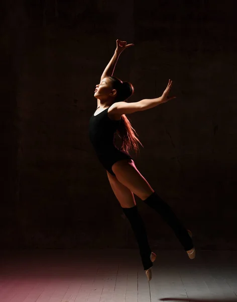 Ung flicka gymnast i svart sport kropp och speciella överdelar hoppa i gymnastiska pose över mörk bakgrund — Stockfoto