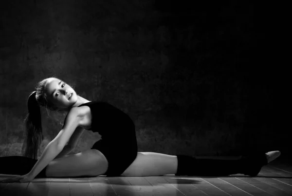 Giovane ragazza sorridente ginnasta con i capelli lunghi in corpo nero sport e tomaie seduti in spago lateralmente su sfondo scuro — Foto Stock
