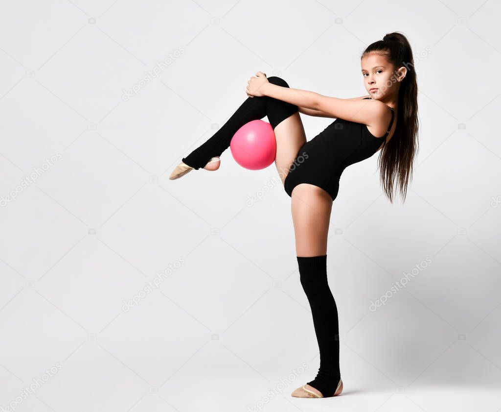 Young girl gymnast in black sport body and uppers standing in bridge pose and holding pink gymnastic ball between leg and neck
