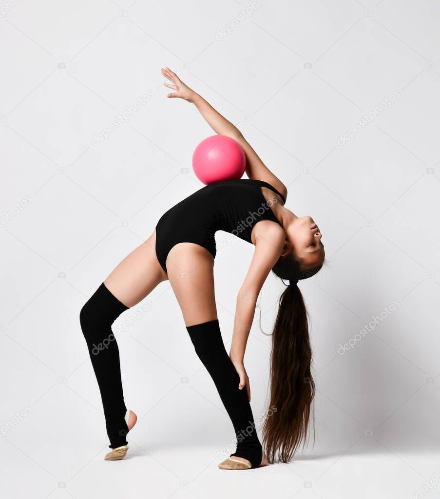 Young girl gymnast in black sport body and uppers staying streched and holding pink gymnastic ball on body over white background