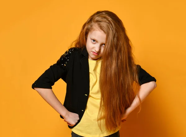 Een tiener in een zwart jasje en een geel t-shirt. Ze ziet er pissig uit, handen op heupen, poserend op oranje achtergrond. Sluiten. — Stockfoto