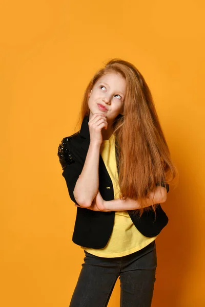 Ginger tienerkind in zwarte jas en broek, geel t-shirt. Ze ziet er attent uit, poserend op oranje achtergrond. Sluiten. — Stockfoto