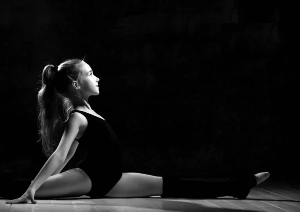 Mladá usměvavá dívka gymnastka s dlouhými vlasy v černém sportovním těle a svršky sedí v motouzu bokem přes tmavé pozadí — Stock fotografie