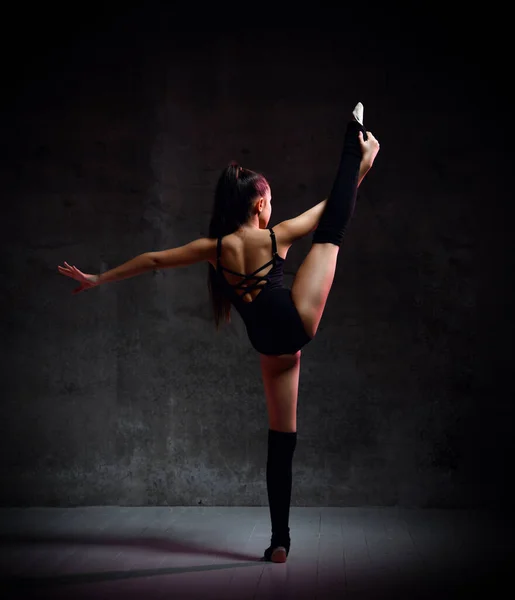 Młoda gimnastyczka w czarnym ciele sportowym i specjalnym obuwiu stojącym tyłem, trzymająca rozciągniętą nogę w dłoni i utrzymująca równowagę — Zdjęcie stockowe