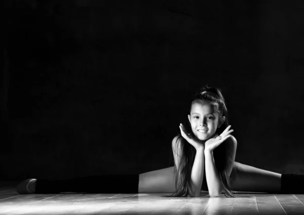Giovane ragazza sorridente ginnasta in corpo nero sport e calzature speciali seduto in spago su sfondo scuro — Foto Stock