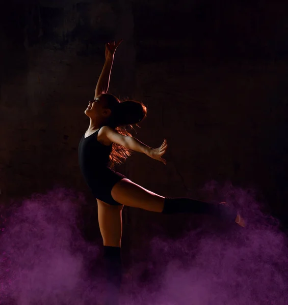 Νεαρό κορίτσι γυμναστής στο μαύρο σώμα του αθλητισμού και το πάνω μέρος κάνοντας κύκλο στο άλμα πάνω από σκούρο φόντο με πολύχρωμα λάμψη — Φωτογραφία Αρχείου