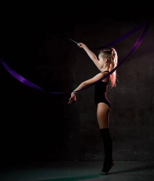 Молодая девушка гимнастка в черном спортивном теле и верхние стоят боком и делают упражнения с синей гимнастической лентой — стоковое фото