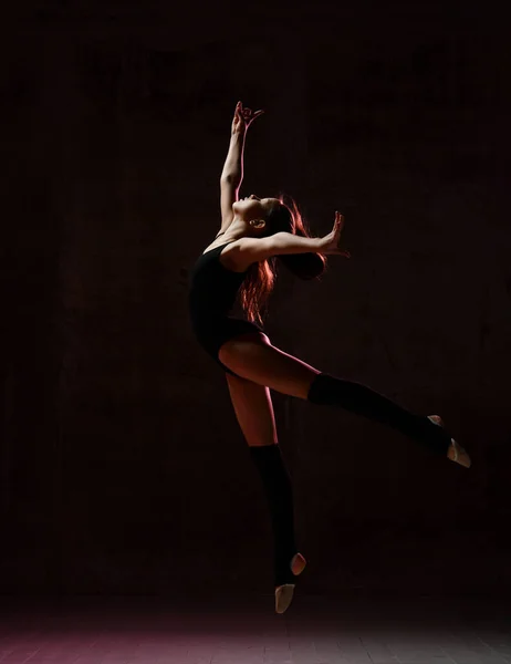 Young girl gymnast in black sport body and special uppers jumping in gymnastic pose over dark background — 스톡 사진