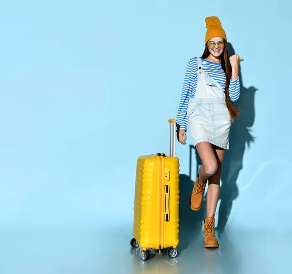 Teenage femelle en jeans dans l'ensemble, sweat-shirt, lunettes de soleil, bottes et chapeau. Souriant, posant avec une valise jaune, fond bleu . — Photo